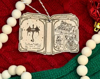 May include: Wooden book-shaped Christmas ornament with etched fantasy illustrations. The ornament features a dragon on one page and a castle scene on the other. It hangs from a twine string and is surrounded by red and green fabric and wooden beads.