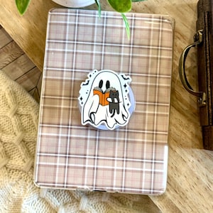 May include: A rectangular e-reader case featuring a beige and white plaid pattern. Centered on the case is a circular pop-out grip decorated with a white ghost illustration holding an orange book and a small black cat, accented by tiny black bats.