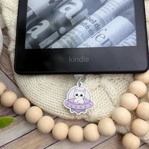 May include: A white cat with pink ears sits in a pink and green spaceship with a white outline. The spaceship is hanging from a black Kindle e-reader.