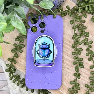 May include: A purple phone case with a speckled design, featuring a pop socket with a blue and purple scarab beetle illustration inside a glass dome. The phone case is resting on a wooden surface, surrounded by green plants.