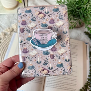 May include: Clear phone case with a pattern of books, coffee cups, eyeglasses, and candles. A sticker on the case features a pink teacup with the words "Coffee, Tea, Books, Cats". The case is held over an open book.