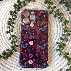 May include: A dark burgundy phone case featuring a floral and butterfly pattern. The design includes pink, purple, and red flowers, along with blue butterflies and spiderwebs. The case has cutouts for the camera and other phone features.
