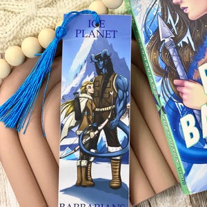 May include: A blue and white bookmark with a blue tassel. The bookmark features an illustration of a blue-skinned humanoid with horns and a woman with blonde hair. The text on the bookmark reads "Ice Planet Barbarians".