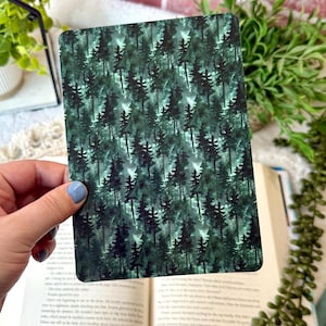 May include: A rectangular card featuring a repeating pattern of dark green pine trees. The trees are set against a lighter green background, creating a forest scene. The card is held over an open book.
