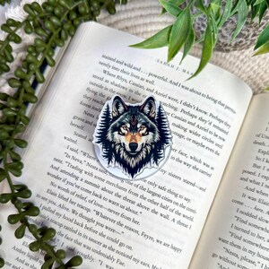 May include: A circular acrylic pin featuring a detailed illustration of a wolf head surrounded by dark pine trees. The wolf has grey, black, and white fur with orange accents on its forehead and nose, set against a background of an open book.
