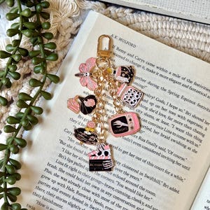 May include: A gold-toned metal keychain featuring multiple enamel charms including a pink moth, a black gramophone, a jar of skulls, a pink television with a skeleton hand and the text COMING, and a black clapperboard with a skeleton hand.