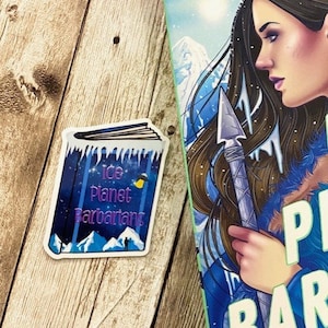 May include: A sticker of a book cover with a blue background and the text "Ice Planet Barbarians". The sticker is on a wooden surface.