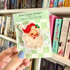May include: A Santa Claus-themed magnetic phone grip from Briar Layne Designs. The grip features a vintage-style illustration of Santa with a red hat, white beard, and a smiling face. The packaging has a green and white checkered pattern.