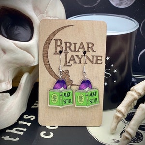 May include: A pair of earrings featuring green book charms with the text "Alien Smut" and purple hair. The earrings are hanging from silver hooks.