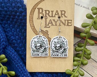 May include: A pair of silver earrings with a black and white design featuring a skull, a book, and the text "Always Fallin' for the Villain" and "Dark Romance Book Club".