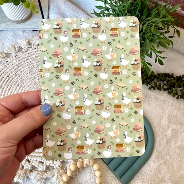 Puede incluir: Un cuaderno con un fondo verde claro que presenta un patrón repetido de gansos blancos, libros y cestas de flores. El cuaderno se sostiene en una mano y tiene un acabado suave y mate.