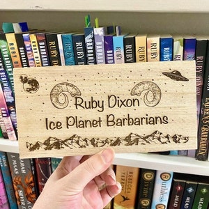 May include: A wooden sign with a laser-engraved design featuring a mountain range, a UFO, and two crescent moons. The text "Ruby Dixon Ice Planet Barbarians" is engraved on the sign.