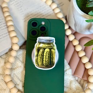 May include: A green phone case with a pickle jar sticker. The sticker features a jar filled with pickles. The phone case is on a white pop socket. The background includes a book, wooden beads, and a plant.