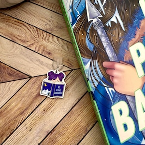Puede incluir: Un charm de libro morado y azul con una niña leyendo un libro titulado "Ice Planet Barbarians".