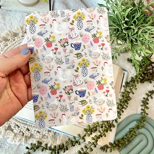 May include: A patterned notebook featuring a whimsical design with chickens, flowers, teacups, and books on a beige background. The notebook is held open, revealing the inside pages. The overall theme is a charming, country-inspired aesthetic.