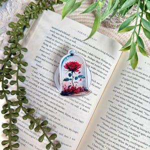 May include: A circular sticker featuring a red rose inside a glass cloche, with scattered red petals at the base. The rose has green leaves and a stem, rendered in a watercolor style. The sticker is placed on an open book page with printed text.