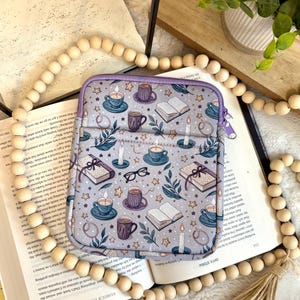 May include: A gray tablet sleeve with a purple zipper, featuring a pattern of books, teacups, candles, stars, and glasses. The sleeve is resting on an open book with wooden beads and greenery in the background.