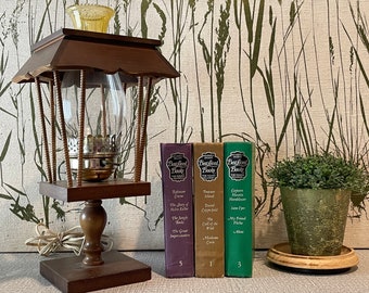 May include: A wooden lamp with a glass shade and a decorative top. The lamp is plugged in and has a cord. The lamp is sitting on a table with three books titled "Best-Loved Books" with different colored covers. The books are in a row and have numbers on the spines. There is a small potted plant to the right of the books.