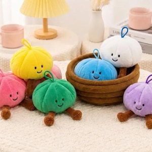 May include: A collection of plush, round, soft toys shaped like vegetables with smiling faces, small brown legs, and a hanging loop on top. The toys come in yellow, pink, green, blue, white, and purple, with some placed inside a brown woven basket.