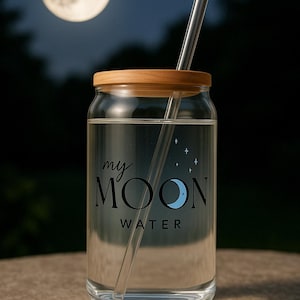 以下が含まれることがあります： 木製の蓋とストローが付いた透明なガラスの水筒。ボトルには「my MOON WATER」という黒と白のデザインが施されています。
