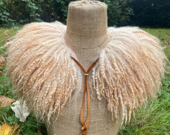 May include: A beige, curly-textured, long-haired sheepskin collar draped over a burlap-covered mannequin torso. The collar features a tan leather cord tie at the center front, which hangs down in two thin, knotted strips against the fabric.