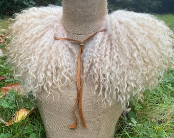 May include: A cream-colored curly wool collar featuring long, soft fibers and a thin brown leather tie cord with a small metallic bead closure. The accessory is displayed on a textured burlap mannequin torso.