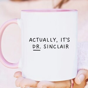 May include: White ceramic mug with a pink rim and handle. The mug has black text that reads "Actually, it's Dr. Sinclair".