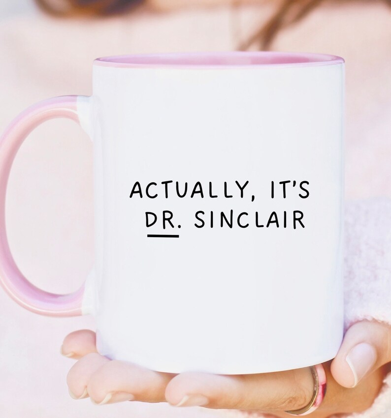 May include: White ceramic mug with a pink rim and handle. The mug has black text that reads "Actually, it's Dr. Sinclair".