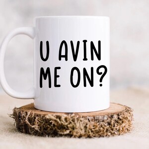 May include: White ceramic mug with the text "U AVIN ME ON?" in black, displayed on a rustic wooden coaster. The mug has a curved handle and is suitable for hot or cold beverages.