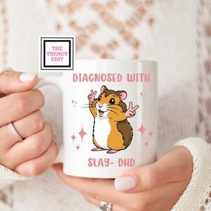 May include: White ceramic mug with a pink and black design. The mug features a cartoon hamster with the text "Diagnosed with Slay-DHD".