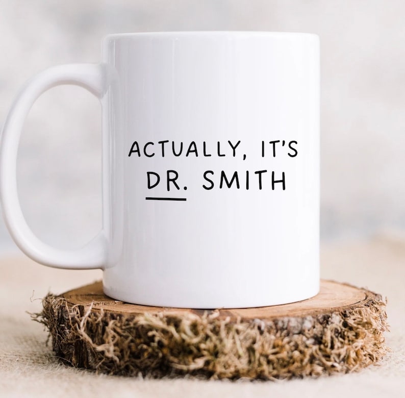 May include: White ceramic coffee mug with black text that reads "Actually, it's Dr. Smith" with a line under the "Dr."