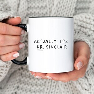 May include: White ceramic mug with a black rim and handle. The mug has black text that reads "Actually, it's Dr. Sinclair" with a line under "Dr."