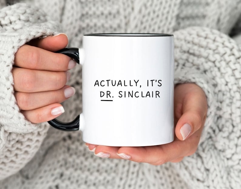 May include: White ceramic mug with a black rim and handle. The mug has black text that reads "Actually, it's Dr. Sinclair" with a line under "Dr."