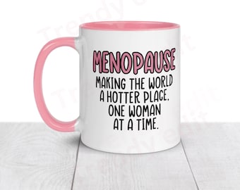 May include: White ceramic mug with a pink interior and handle. The mug features the text "MENOPAUSE MAKING THE WORLD A HOTTER PLACE. ONE WOMAN AT A TIME." in black and pink lettering. A humorous gift.