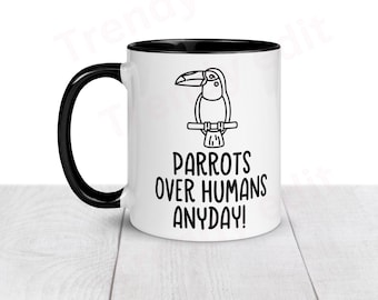 May include: White ceramic coffee mug with a black rim and handle. The mug has a black and white illustration of a toucan and the text "Parrots over humans anyday!"