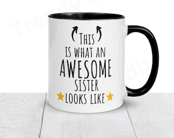 May include: White ceramic mug with a black rim and handle. The mug has black text that reads "(This) is what an awesome sister looks like" with two gold stars on either side of the text.