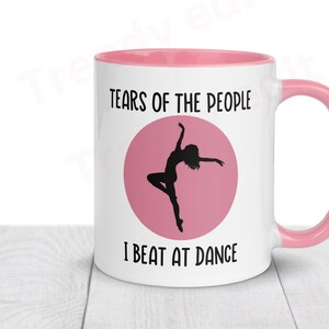 May include: White ceramic mug with a pink rim and handle. The mug has a pink circle with a silhouette of a dancer and the text "Tears of the people I beat at dance".