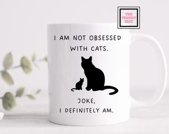 May include: White ceramic coffee mug with a black silhouette of two cats and the text "I am not obsessed with cats. Joke, I definitely am."