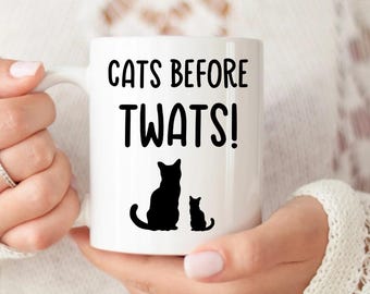 May include: White ceramic coffee mug with black text that reads "Cats Before Twats!" and a black silhouette of two cats.
