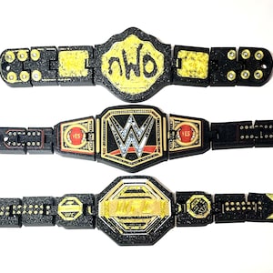 May include: Three toy championship belts are shown against a white backdrop. The belts are black with gold and yellow detailing. One belt features "nWo" in yellow, another has a "W" logo, and the third has "UFC" and "JKM" in yellow. A coin is next to the belts.