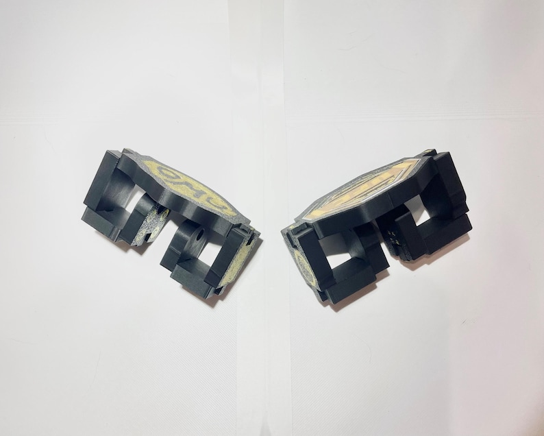May include: Two black, geometric objects with a gold design. The objects appear to be made of plastic or a similar material. The design on each object is circular and has a yellow and gold colour scheme.