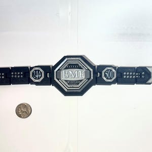 May include: A black and silver championship belt with the text "BMF" in the center. The belt has the numbers "244" and "500" on either side. The belt is lying on a white surface.