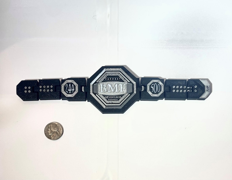 May include: A black and silver championship belt with the text "BMF" in the center. The belt has the numbers "244" and "500" on either side. The belt is lying on a white surface.