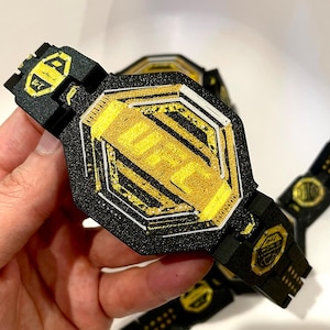 May include: A black and gold UFC championship belt with the UFC logo in the centre. The belt has a black strap and gold accents. The belt is held in a person's hand.