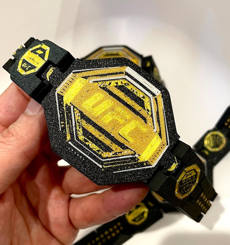 May include: A black and gold UFC championship belt with the UFC logo in the centre. The belt has a black strap and gold accents. The belt is held in a person's hand.