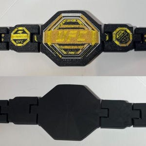May include: A black and gold UFC championship belt. The belt has a central gold emblem with the UFC logo and text, framed by a black border. The strap is segmented and black, with more UFC logos.