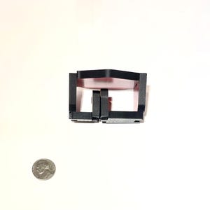 May include: A black plastic component with a rectangular design and a central aperture. A coin is placed beside the object for scale. The object is set against a white backdrop. The coin is approximately 2.4 cm in diameter.