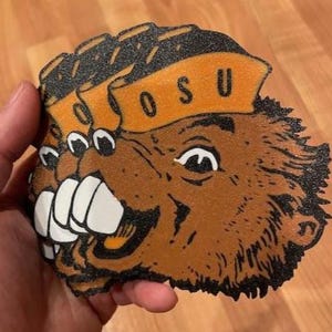 May include: A brown and orange beaver-shaped item with the letters "OSU" on a banner. The beaver has large white teeth and is held in a hand. The item is likely a coaster or decorative piece.