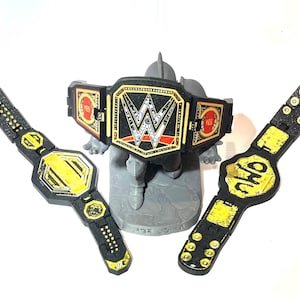 May include: Three toy wrestling championship belts. The belts are black and gold, with the WWE logo on the center belt. The other two belts have yellow accents and the letters "NWO" on one. The belts are on a grey stand.