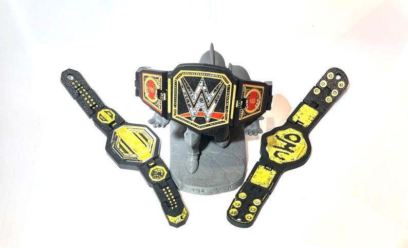 May include: Three toy wrestling championship belts. The belts are black and gold, with the WWE logo on the center belt. The other two belts have yellow accents and the letters "NWO" on one. The belts are on a grey stand.